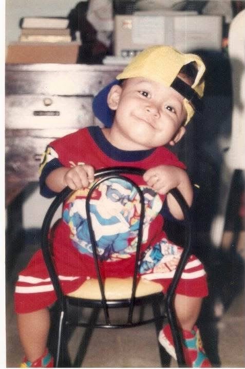 Reva as a toddler, sitting on a chair, grinning under an oversized cap