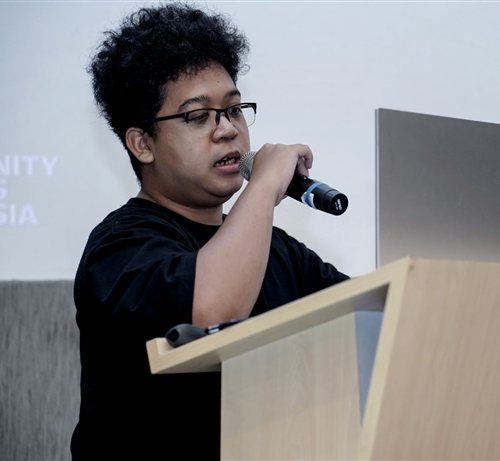 Reva delivering a talk at a podium, microphone in hand, in front of a presentation screen